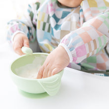Load image into Gallery viewer, Close-up of a child wearing a Bumkins Sleeved Bib: Motley Check reaching into a green bowl while holding a spoon.
