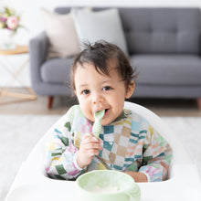 Load image into Gallery viewer, A toddler wears a Bumkins Sleeved Bib: Motley Check while sitting in a high chair, chewing a green spoon near a green bowl.

