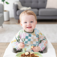 Load image into Gallery viewer, A smiling toddler in a high chair wears a Bumkins Sleeved Bib: Motley Check, holding a spoon with waffles and bananas on a plate in front.
