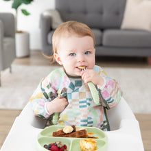 Load image into Gallery viewer, A toddler in a Bumkins Sleeved Bib: Motley Check sits in a high chair, eating fruit and waffles with a fork from a green plate.
