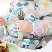 Load image into Gallery viewer, Close-up of a child in a colorful checkered sleeved bib holding Bumkins Spoon + Fork: Sage.
