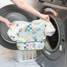 Load image into Gallery viewer, A person holds Bumkins Sleeved Bib: Motley Check in front of an open front-loading washing machine.
