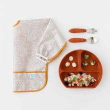 Load image into Gallery viewer, Bumkins Sleeved Bib: Wander is folded on a white background, next to a dark orange divided plate of food and utensils.
