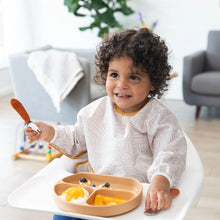 Load image into Gallery viewer, A curly-haired toddler grins in a high chair, clutching a spoon and wearing a Bumkins Sleeved Bib: Wander as they eat from a tan dish.

