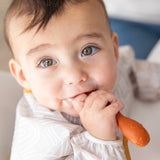 A bright-eyed baby wearing a beige bib chews an orange Bumkins Silicone Dipping Spoons 3 Pack: Rocky Road.