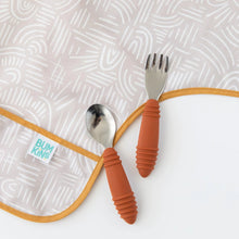 Load image into Gallery viewer, Close-up of a spoon and fork with orange rubber handles on top of a Bumkins Sleeved Bib: Wander on a white background.
