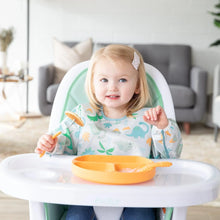 Load image into Gallery viewer, A toddler in a high chair, wearing a Bumkins Sleeved Bib: Dinosaurs holds a fork with a pancake near an orange plate.
