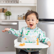 Load image into Gallery viewer, A joyful toddler in a high chair, wearing a Bumkins Sleeved Bib: Dinosaurs, raises their arm excitedly, with orange utensils on the tray.

