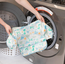 Load image into Gallery viewer, A person holds Bumkins Sleeved Bib: Dinosaurs in front of an open front-loading washing machine.
