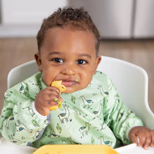 Load image into Gallery viewer, A baby in a high chair, wearing a Bumkins Sleeved Bib: Llamas, chews on a yellow silicone spoon with a yellow plate in front.
