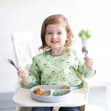 Load image into Gallery viewer, A toddler wearing a Bumkins Sleeved Bib: Llamas in a high chair happily holds a spoon and fork over a gray divided plate.
