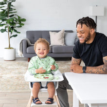 Load image into Gallery viewer, A joyful toddler enjoys mealtime in a Bumkins Sleeved Bib: Llamas, as a tattooed man sitting at a white table smiles and watches.
