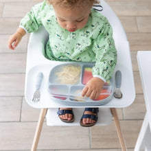 Load image into Gallery viewer, A toddler in a Bumkins Sleeved Bib: Llamas sits in a high chair, removing a transparent lid from a gray divided plate with utensils nearby.
