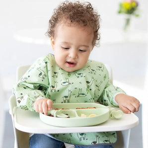 A child in a high chair wearing a Bumkins Sleeved Bib: Llamas eats from a green divided plate with toddler utensils.