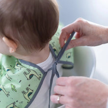 Load image into Gallery viewer, Adult hands tie the ribbon closure of a Bumkins Sleeved Bib: Llamas around a child's neck.
