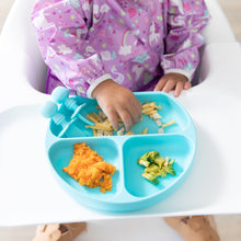 Load image into Gallery viewer, A baby in a Bumkins Sleeved Bib: Unicorns enjoys sweet potato, avocado, and macaroni from a blue plate on a highchair.

