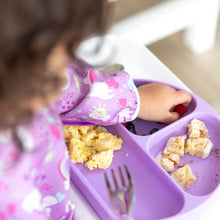 Load image into Gallery viewer, A child in a Bumkins Sleeved Bib: Unicorns enjoys bread, berries, and eggs from a purple divided plate.
