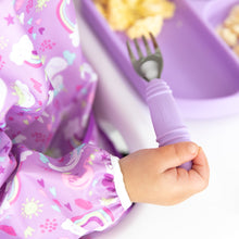 Load image into Gallery viewer, Close-up of a child in a purple unicorn-patterned bib holding a Bumkins Spoon + Fork: Lavender over a matching plate with eggs.
