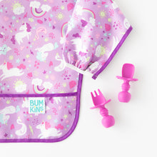 Load image into Gallery viewer, Close-up of Bumkins Sleeved Bib: Unicorns on a white background next to magenta toddler utensils.
