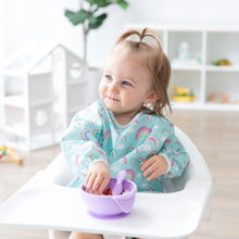 Load image into Gallery viewer, A toddler in a high chair grins while eating from a purple bowl, wearing a Bumkins Sleeved Bib: Rainbows in a bright room.
