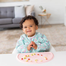 Load image into Gallery viewer, A curly-haired toddler eats fruit from a pink silicone mat, wearing a Bumkins Sleeved Bib: Rainbows in a bright living room.
