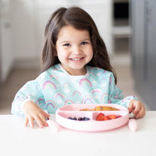 Load image into Gallery viewer, A happy girl in a Bumkins Sleeved Bib: Rainbows enjoys a meal from a pink plate at a white table.
