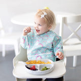 A toddler in a high chair, wearing a Bumkins Sleeved Bib: Rainbows, happily enjoys pasta, strawberries, and broccoli from a gray plate.