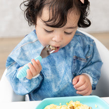 Load image into Gallery viewer, A toddler with curly hair eats scrambled eggs in a high chair, wearing a Bumkins Sleeved Bib: Vintage Denim.
