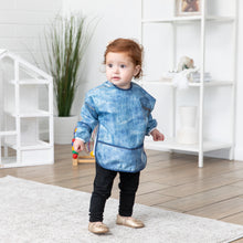 Load image into Gallery viewer, A red-haired child stands in a bright room, wearing a Bumkins Sleeved Bib: Vintage Denim.
