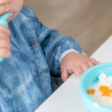 Load image into Gallery viewer, Close-up of a child in a Bumkins Sleeved Bib: Vintage Denim at a white table with a blue bowl of white food; focus is on the elastic cuff.
