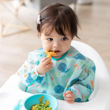 Load image into Gallery viewer, A toddler in a Bumkins Sleeved Bib: Ocean Life sits in a high chair, chewing an orange spoon with a blue bowl on the tray.
