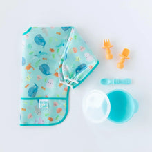 Load image into Gallery viewer, Bumkins Sleeved Bib: Ocean Life lays folded on a white background next to orange baby utensils and a blue bowl and spoon set.
