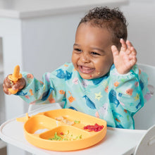 Load image into Gallery viewer, A baby wearing a Bumkins Sleeved Bib: Ocean Life smiles in a high chair, eating from an orange divided plate.
