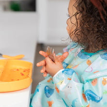 Load image into Gallery viewer, A curly-haired child in a Bumkins Sleeved Bib: Ocean Life holds a messy fork next to an orange plate with food remnants.
