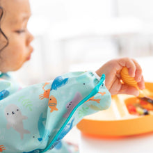 Load image into Gallery viewer, Close-up of a child in a Bumkins Sleeved Bib: Ocean Life, their arm and face visible as they use an orange spoon to eat from an orange plate.
