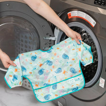Load image into Gallery viewer, A person holds Bumkins Sleeved Bib: Ocean Life in front of an open front-loading washing machine.

