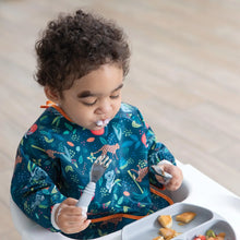 Load image into Gallery viewer, A curly-haired toddler in a Bumkins Sleeved Bib: Jungle sits in a high chair, holding a fork and spoon and looking down at a gray plate.
