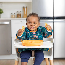 Load image into Gallery viewer, A smiling toddler in a high chair, wearing Bumkins Sleeved Bib: Jungle, holds orange utensils near an orange divided plate.
