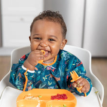 Load image into Gallery viewer, A happy baby in a high chair wears a Bumkins Sleeved Bib: Jungle while chewing an orange baby; an orange plate of food sits on the tray.
