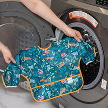 Load image into Gallery viewer, A person holds Bumkins Sleeved Bib: Jungle in front of an open front-loading washing machine.
