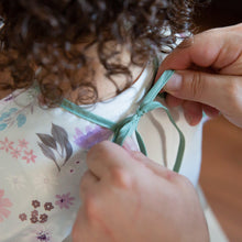 Load image into Gallery viewer, Hands tying the ribbon closure of a Bumkins Sleeved Bib: Floral around a child's neck.

