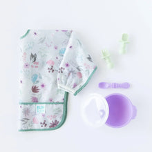 Load image into Gallery viewer, Bumkins Sleeved Bib: Floral lays folded on a white background next to a purple bowl and spoon set with lid, and green toddler utensils.
