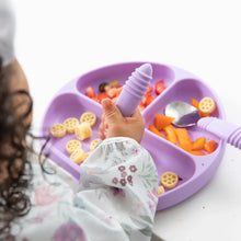 Load image into Gallery viewer, Close-up of a child in a Bumkins Sleeved Bib: Floral enjoying colorful foods from a purple divided plate with utensils at a white table.
