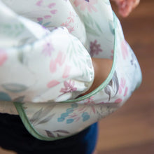 Load image into Gallery viewer, Close-up of a child wearing a Bumkins Sleeved Bib: Floral, made from waterproof fabric, with their hand tucked in the front pocket.
