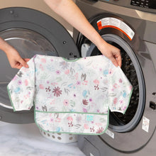 Load image into Gallery viewer, A person holds Bumkins Sleeved Bib: Floral in front of an open front-loading washing machine.
