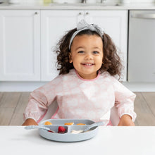 Load image into Gallery viewer, A young girl with curly hair and gray bow wears a Bumkins Sleeved Bib: Lace, sitting at a white table with fruit on a gray divided plate.
