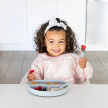 Load image into Gallery viewer, A curly-haired toddler in a Bumkins Sleeved Bib: Lace smiles, holding a fork with fruit, seated at a table with a divided plate of food.
