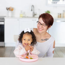 Load image into Gallery viewer, A woman in glasses watches a child in a Bumkins Sleeved Bib: Lace eat from a pink plate using a fork at a white table.
