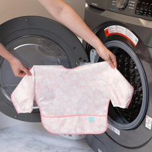 Load image into Gallery viewer, A person holds Bumkins Sleeved Bib: Lace in front of an open front-loading washing machine.
