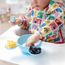 Load image into Gallery viewer, A child wearing Bumkins Sleeved Bib: Mickey &amp; Friends Check Mates eats from a blue bowl with cheese and blueberries on a highchair.

